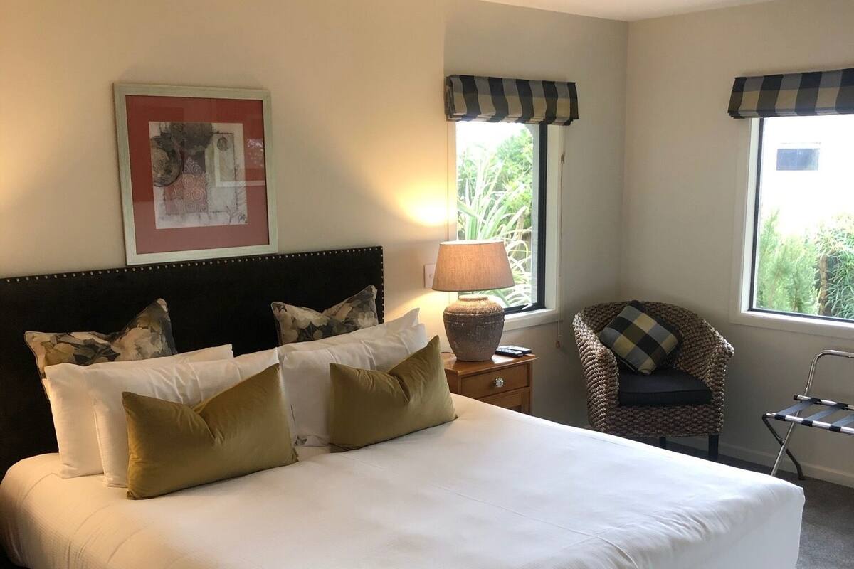 A neatly made bed with white linens and multiple pillows, a framed artwork on the wall, a window with a view of greenery, and a wicker chair with a checkered pillow.