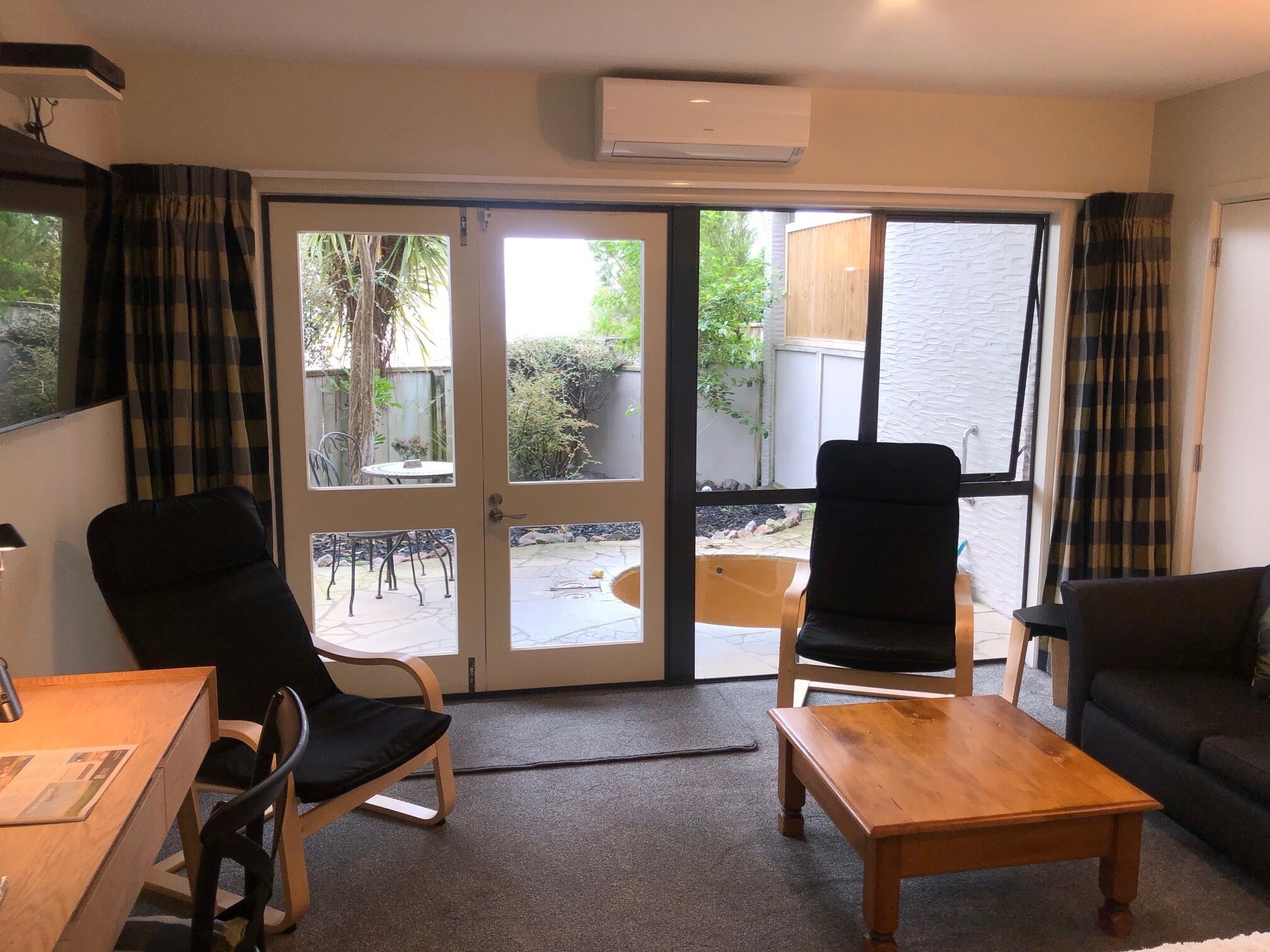 Una sala de estar con un sofá, dos sillones, una mesa de centro de madera y una mesa de comedor con sillas. Hay puertas de cristal grandes que dan a un patio exterior con una mesa y sillas.