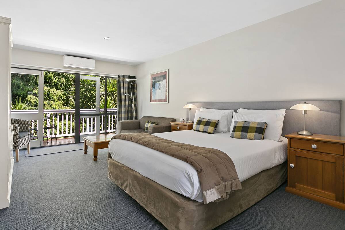 A spacious bedroom with a large bed, a nightstand, a wooden cabinet, and a balcony with a view of greenery.