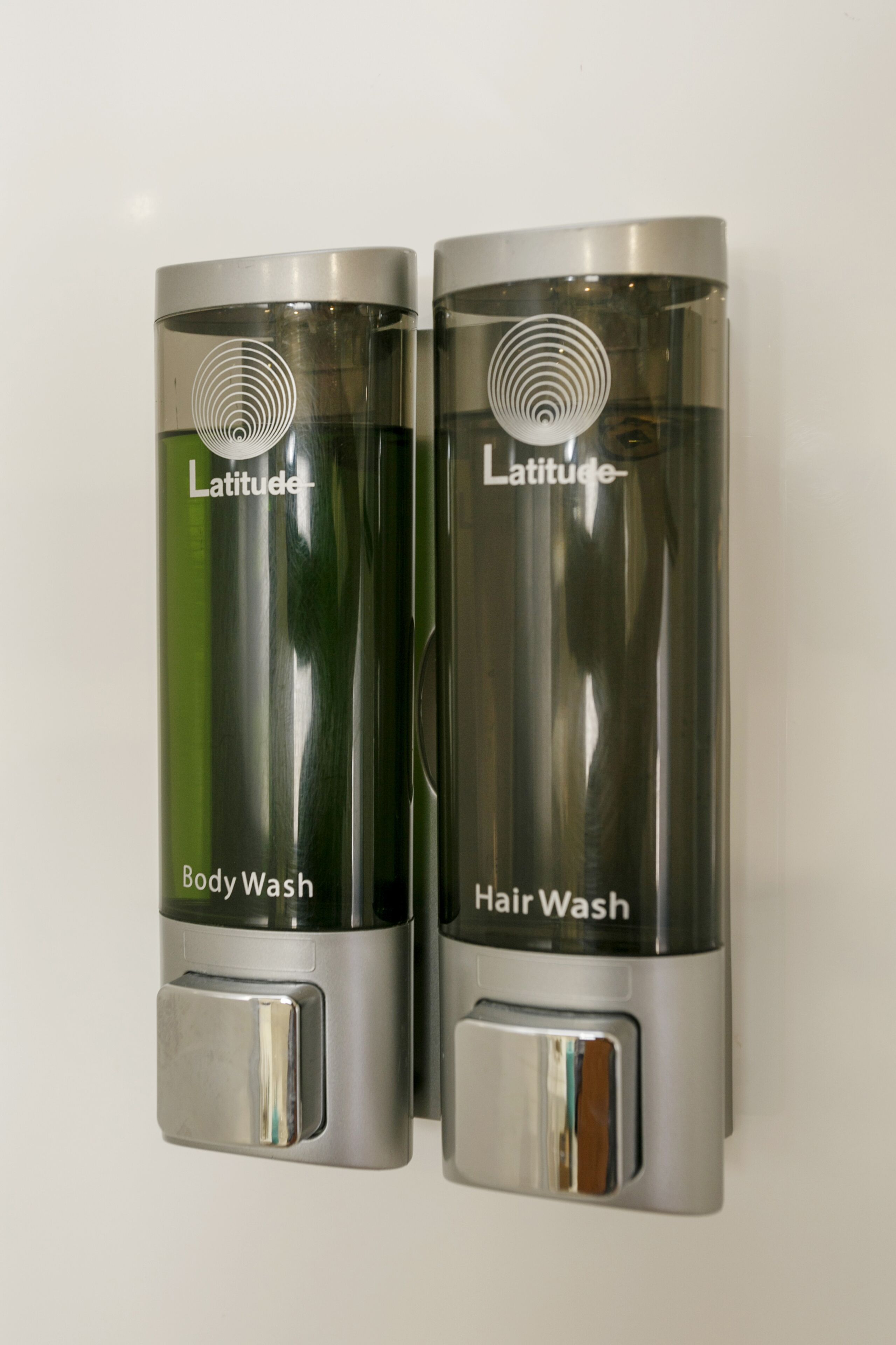 Dos dispensadores de acero inoxidable con las etiquetas 'Latitude Body Wash' y 'Latitude Hair Wash' montados en una pared.