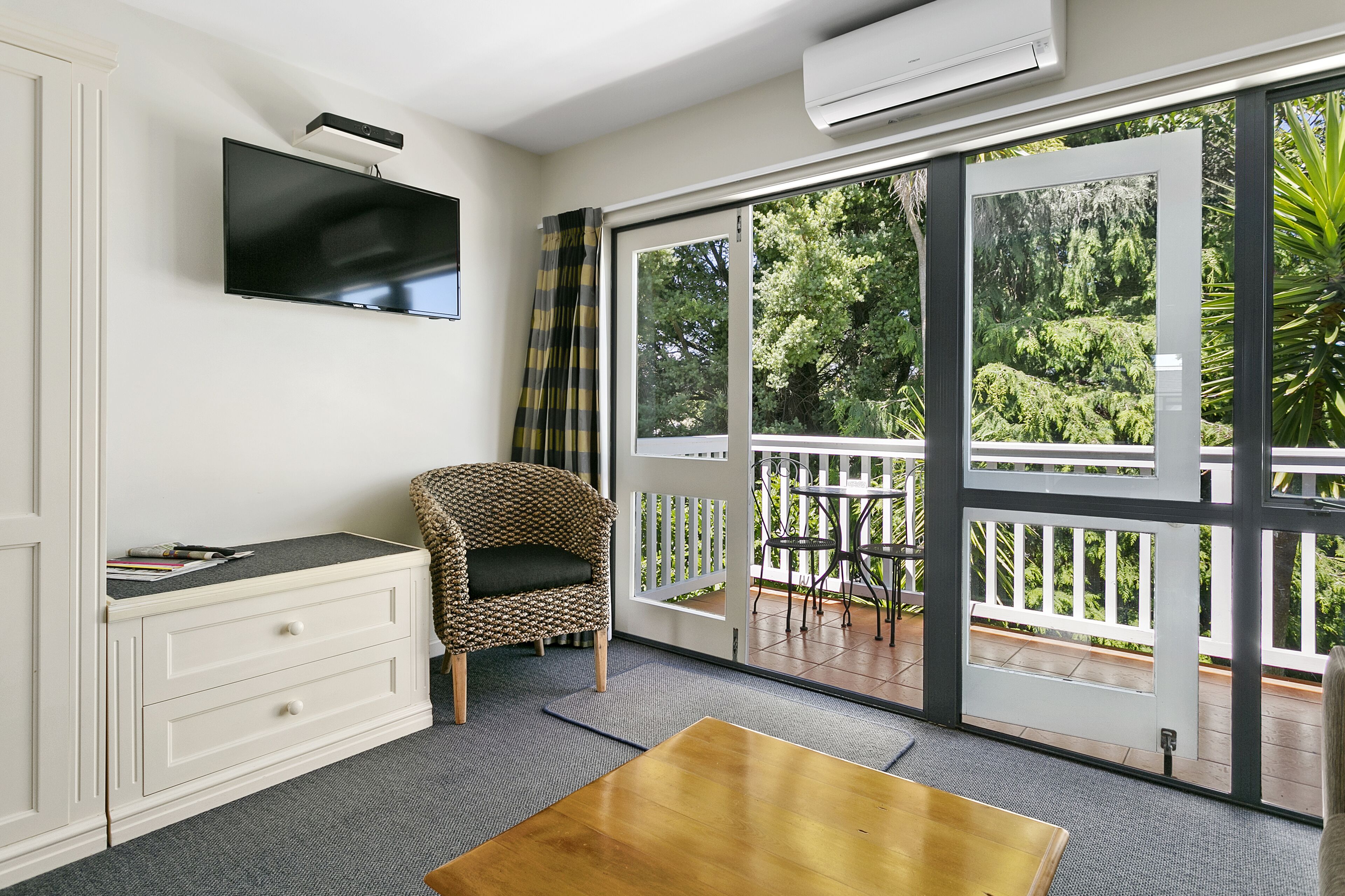 A room with a flat-screen TV mounted on the wall, a wicker chair with a cushion, a wooden table, and a sliding glass door leading to an outdoor deck with a white railing and greenery.