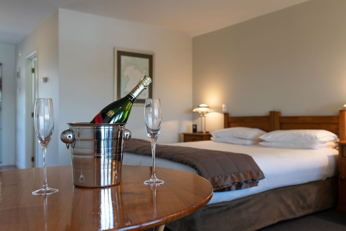 Habitación de hotel con cama, mesita de noche, lámpara y una botella de champán en un cubo con copas.