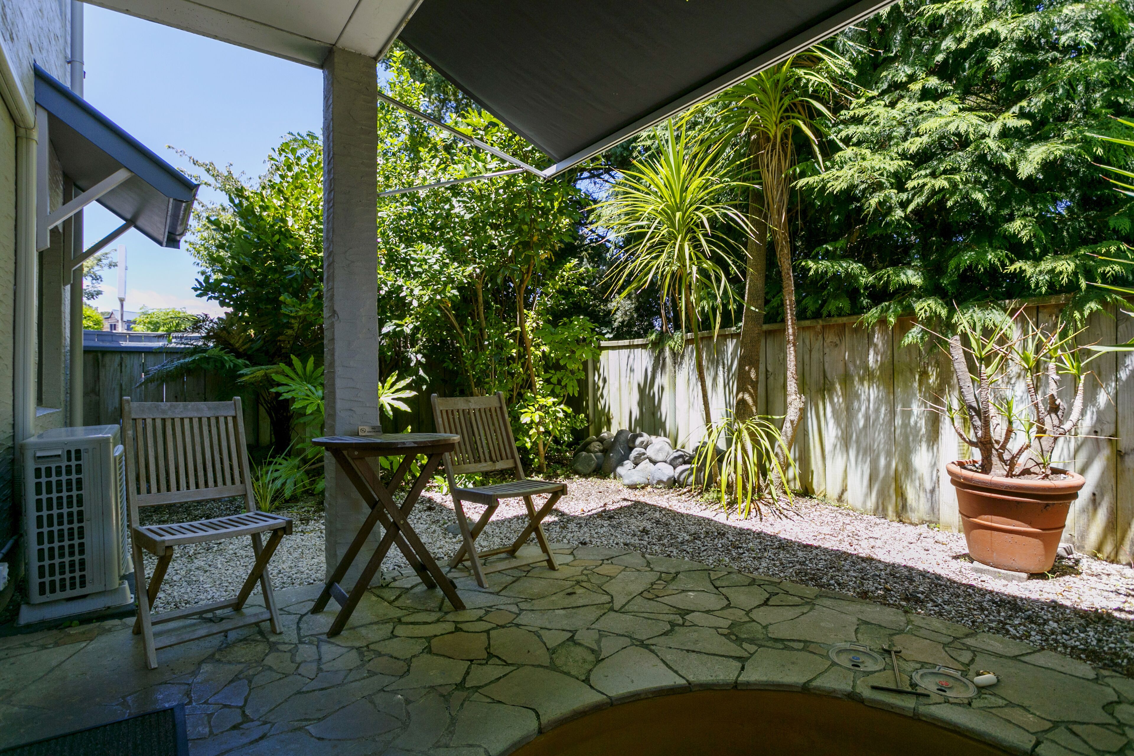 Um pátio ao ar livre com piso de pedra, uma mesa de madeira, duas cadeiras de madeira, um vaso de planta e uma pequena piscina.