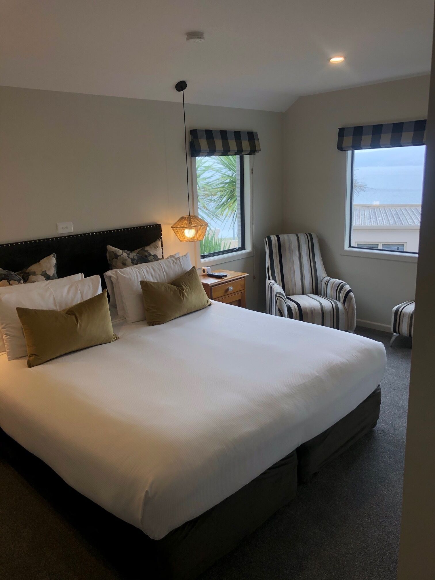 A large bed with multiple pillows, a bedside table with a lamp, a striped armchair, and a window with a view of the sea.