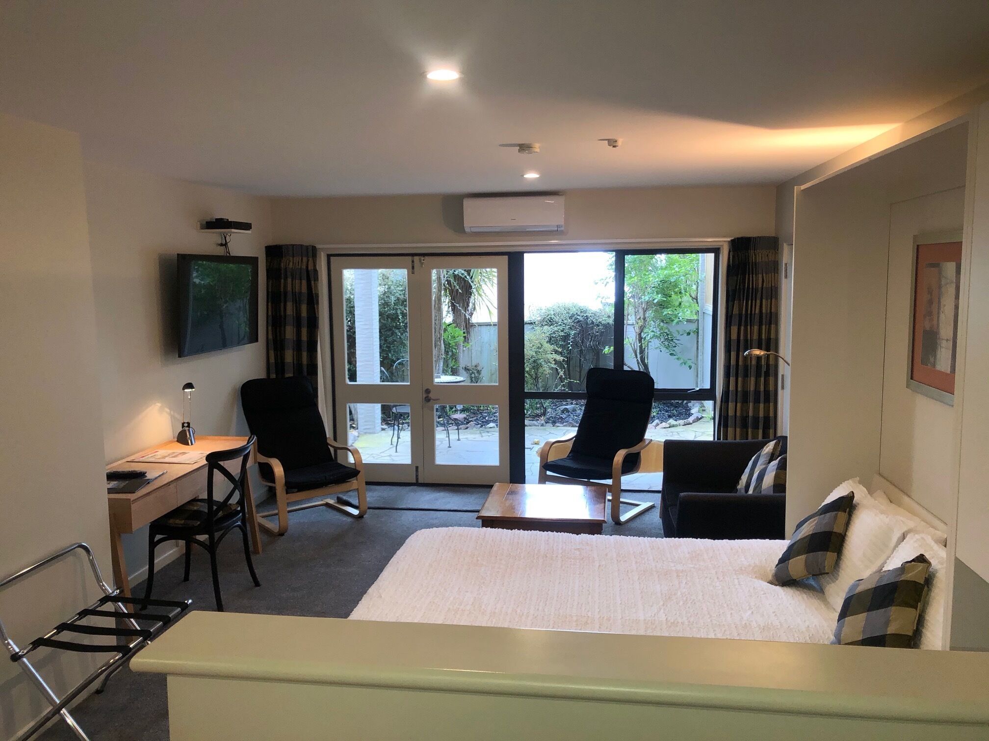 Una habitación de hotel con una cama, dos sillones, un escritorio con lámpara, un televisor y una puerta corrediza de vidrio que da a un balcón.