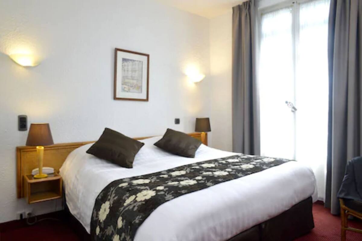 Une chambre d’hôtel avec un lit, deux lampes de chevet, une table de nuit, un tableau encadré au mur et une fenêtre avec des rideaux.
