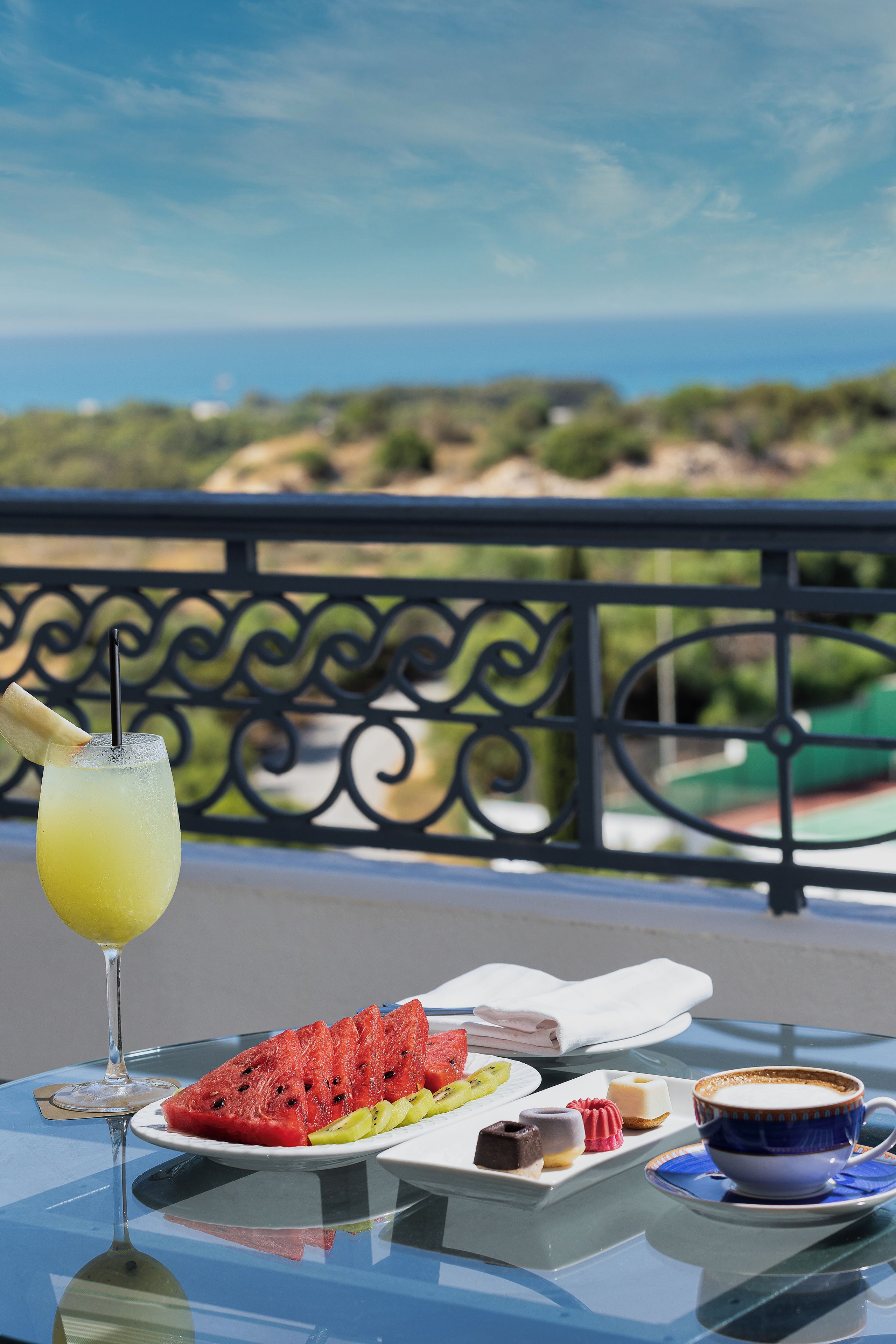 A table with a glass of drink, a plate of food, and a cup on a balcony overlooking a scenic view.