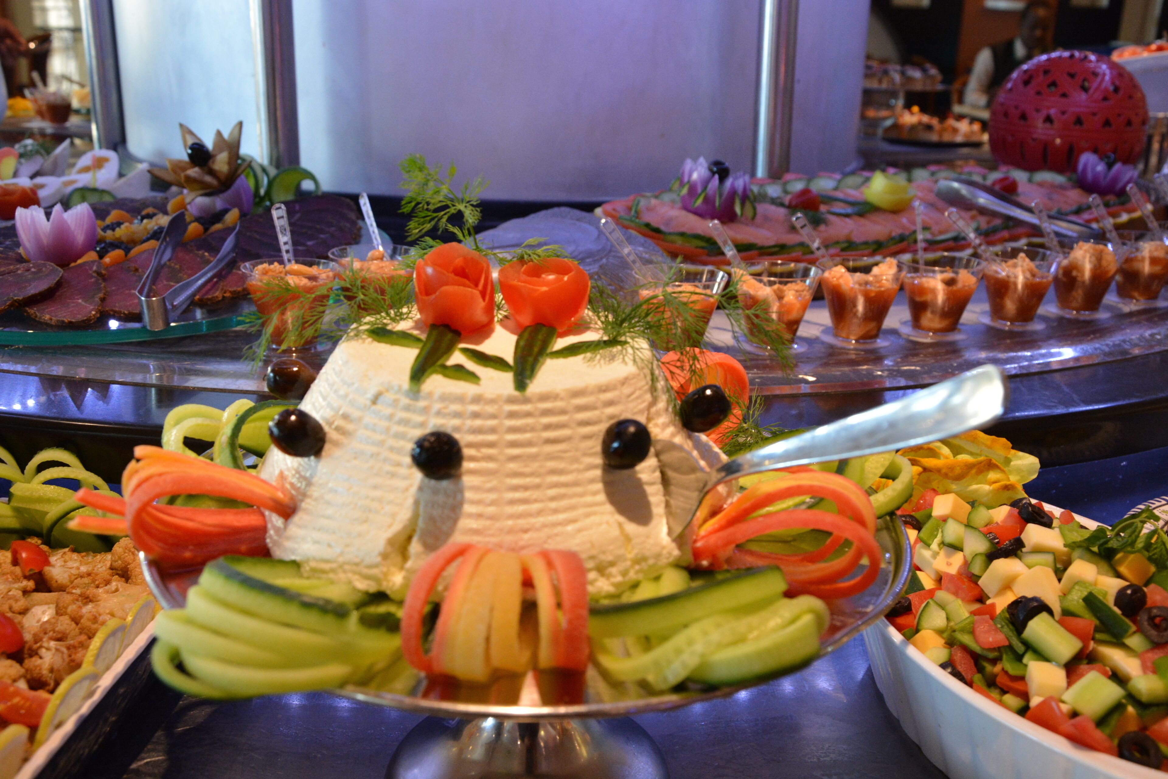 Ein reichhaltiges Buffet mit verschiedenen Speisen, darunter eine tortenartige Gesichtskreation, umgeben von Gemüse und weiteren Gerichten.