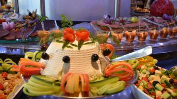 Ein reichhaltiges Buffet mit verschiedenen Speisen, darunter eine tortenartige Gesichtskreation, umgeben von Gemüse und weiteren Gerichten.