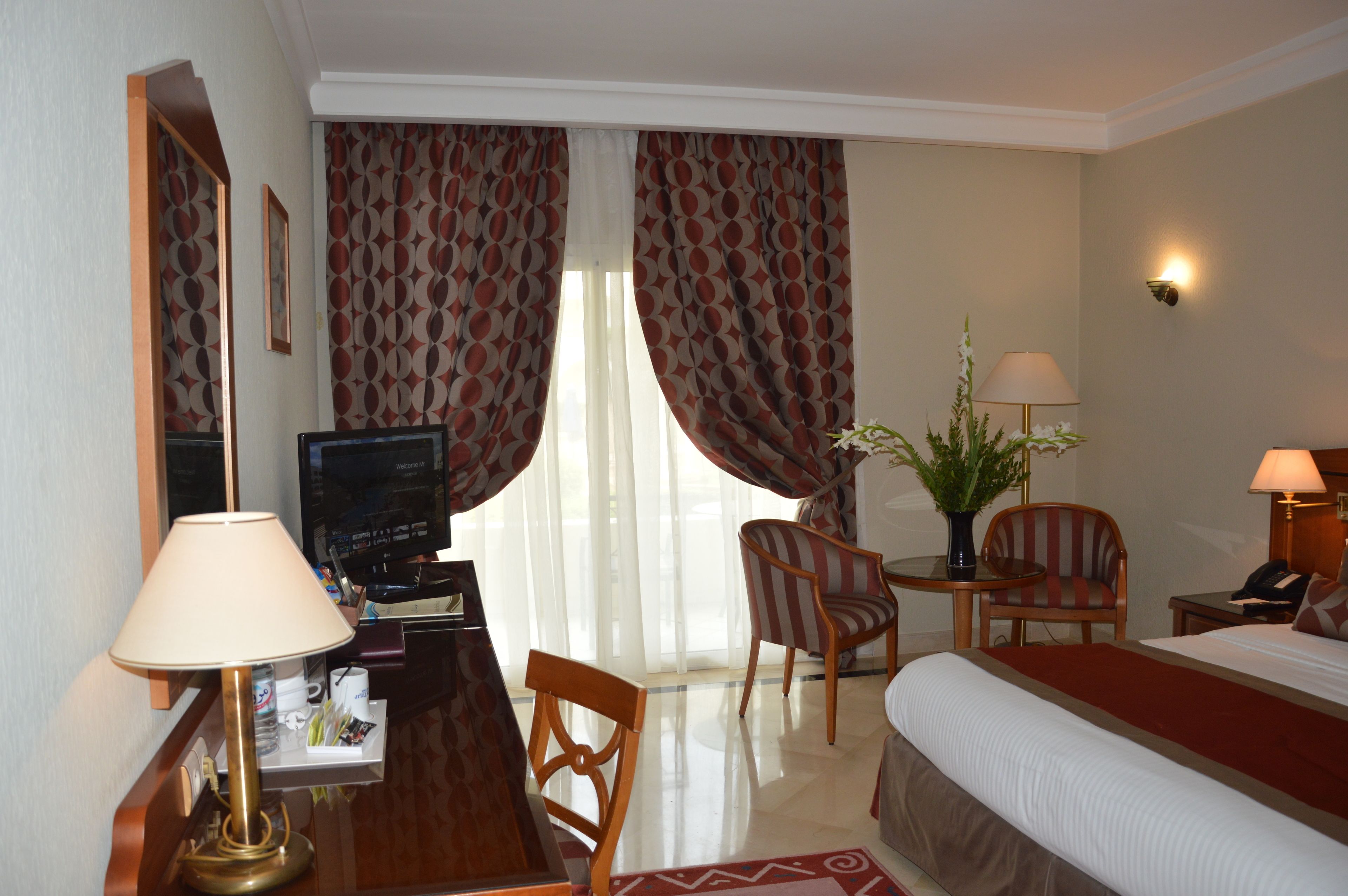 A hotel room with a bed, a desk with a lamp, a television, two chairs, a small table with a plant, and a nightstand with a lamp.
