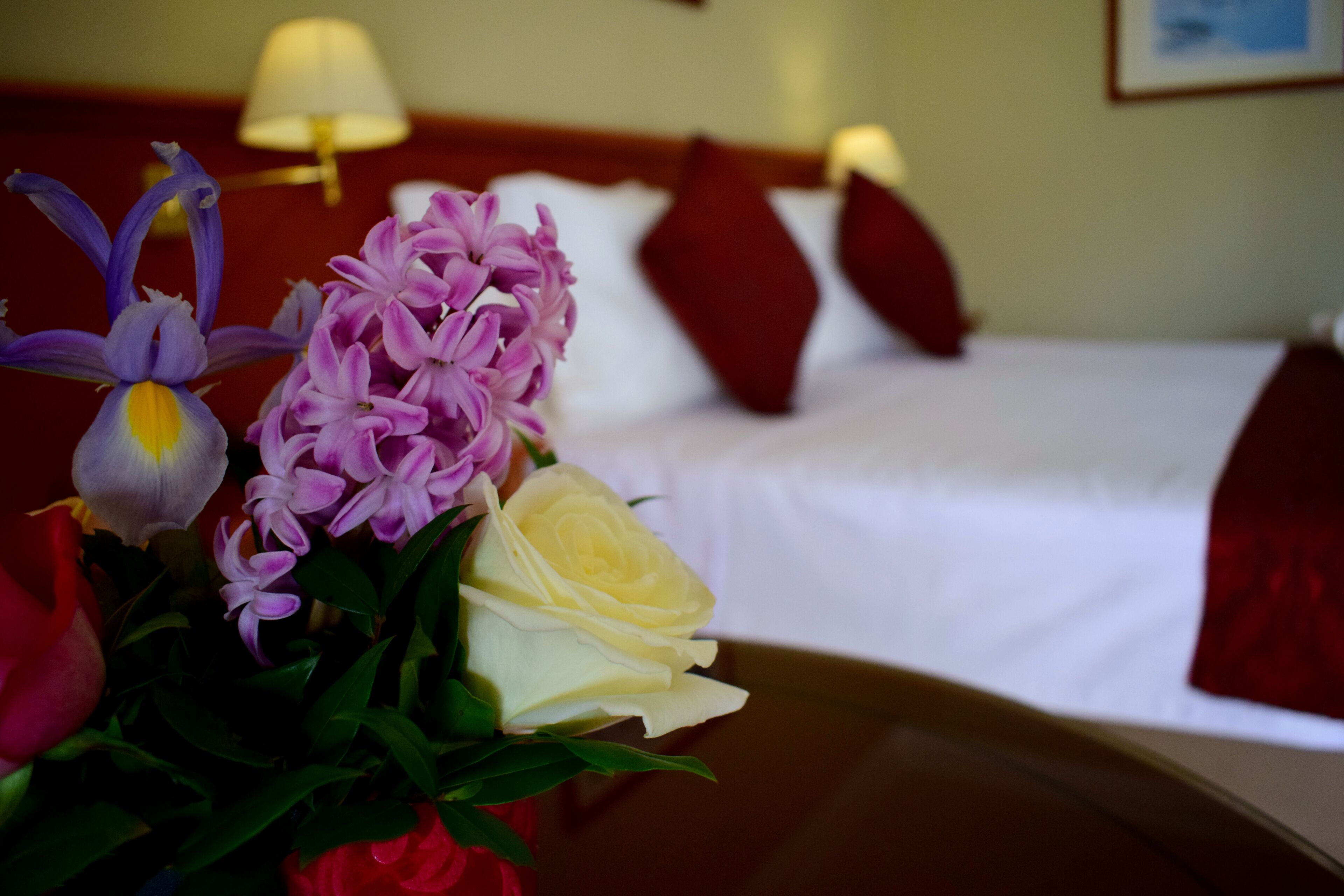 A hotel room with a bed, pillows, and a floral arrangement on the bedside table.