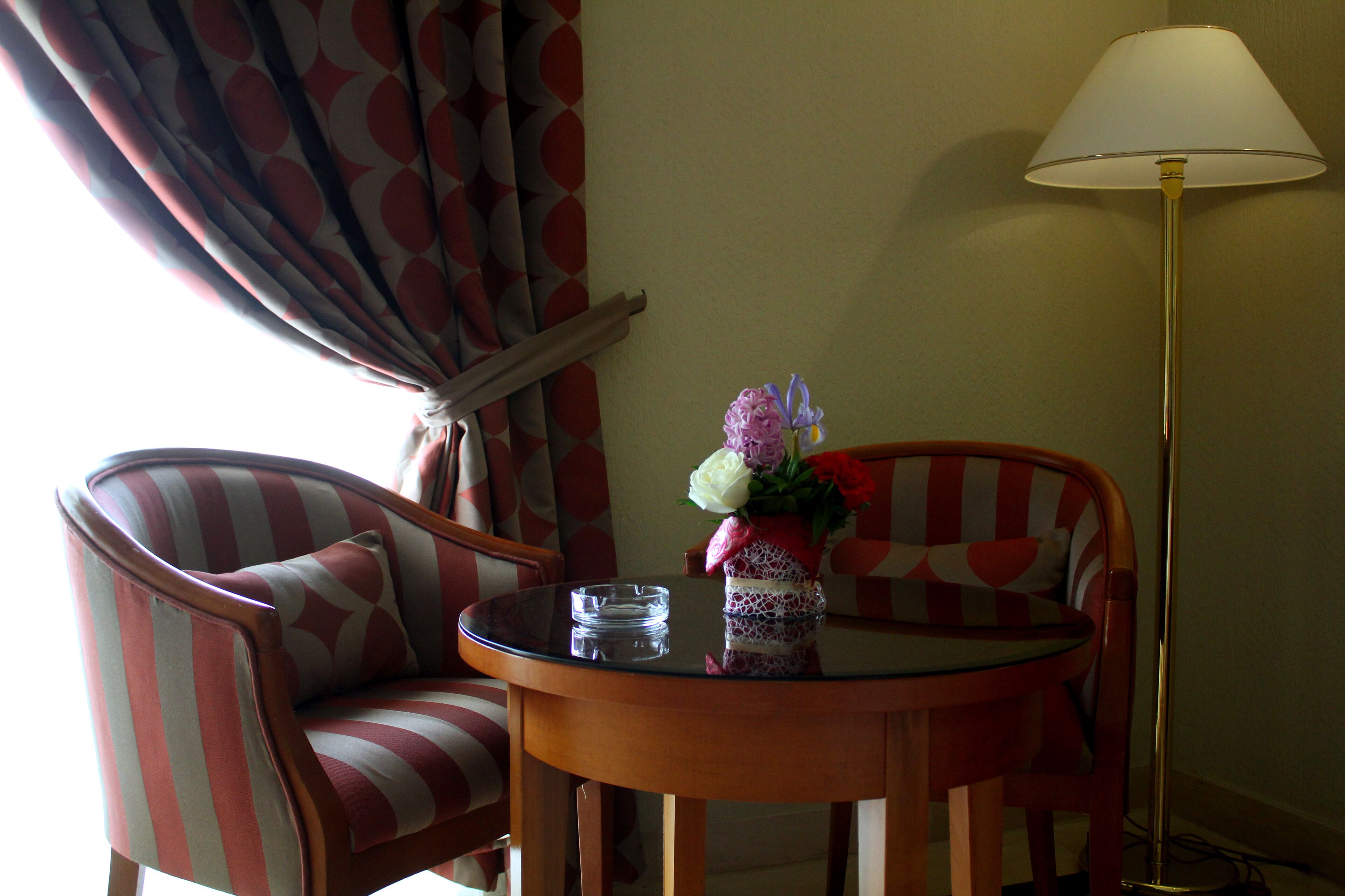 A room with a round glass table, two striped armchairs, a vase with flowers, and a standing floor lamp.
