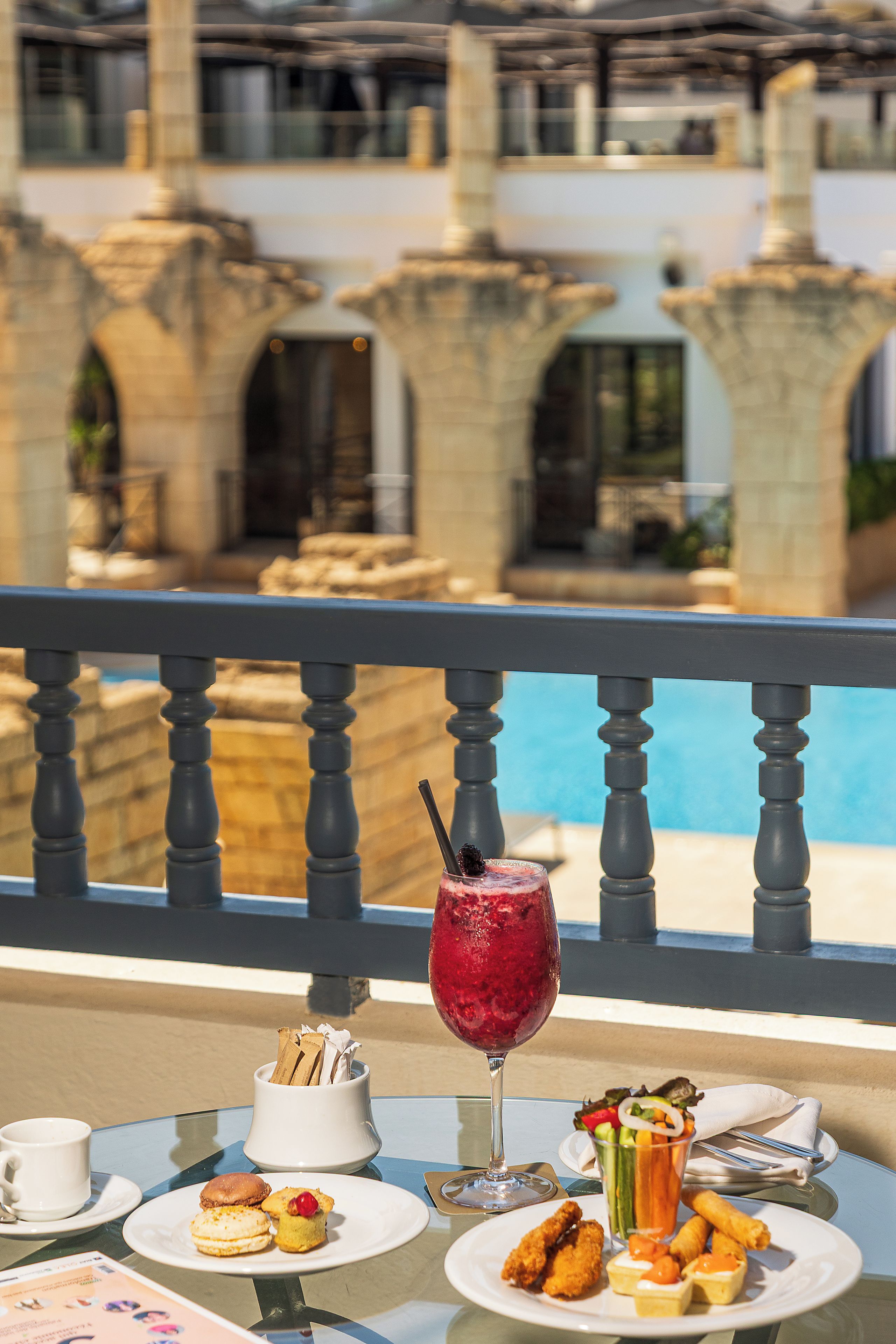 A dining table with a variety of food items and a glass of beverage, overlooking a pool area with stone pillars and a clear sky.