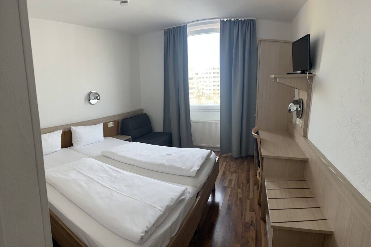 Et hotelværelse med to senge, et skrivebord, en stol, et fjernsyn og et vindue med gardiner.