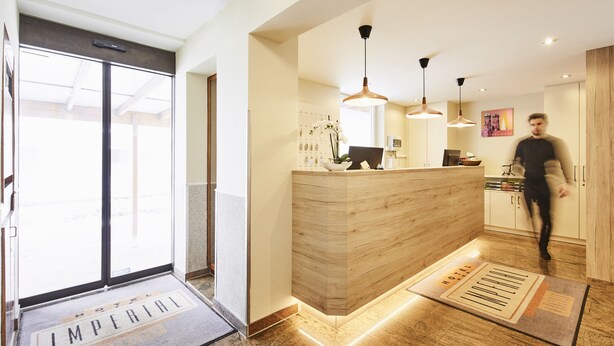 A hotel reception area with a wooden reception desk, pendant lighting, and a view of the outside through a glass door. - Hotel Imperial (Munich)
