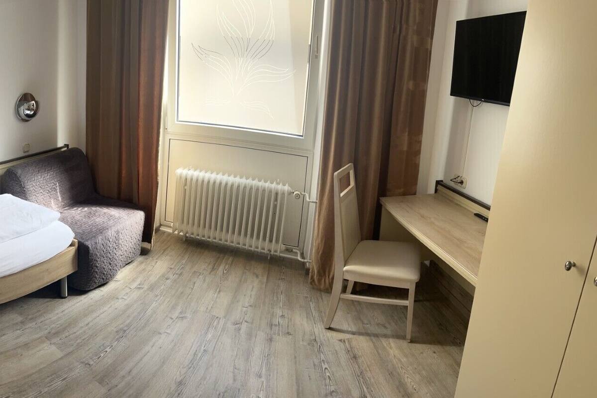 Et hotelværelse med seng, stol, skrivebord, radiator og fjernsyn.