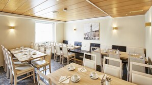 A modern dining area with wooden tables and white chairs, a large window, and a cityscape painting on the wall. - Hotel Imperial (Munich)