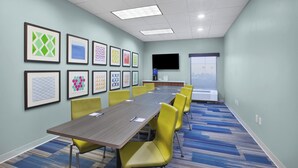 A conference room with a long table, yellow chairs, framed artwork on the walls, a flat-screen TV, and a large window.