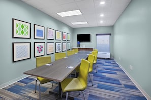 A conference room with a long table, yellow chairs, framed artwork on the walls, a flat-screen TV, and a large window.