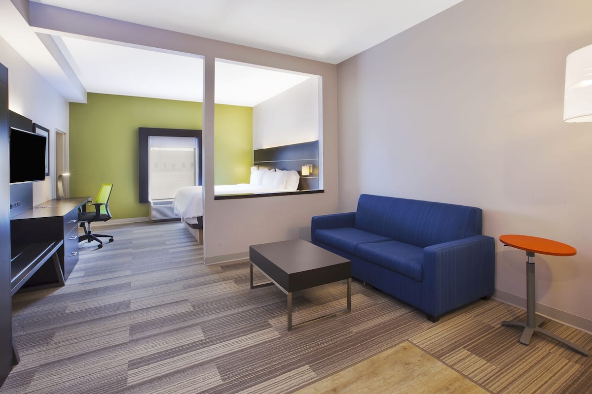 A modern hotel room with a blue sofa, a small table, a TV, a desk with a chair, and a view of a balcony.