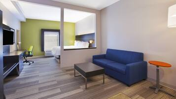 Et moderne hotelværelse med blå sofa, et lille bord, et fjernsyn, et skrivebord med stol og balkonudsigt.