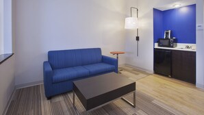 A modern living room with a blue sofa, a dark coffee table, and a small round table. A kitchen area with a microwave and a sink is visible in the background. - Holiday Inn Express Tiffin by IHG (Tiffin)