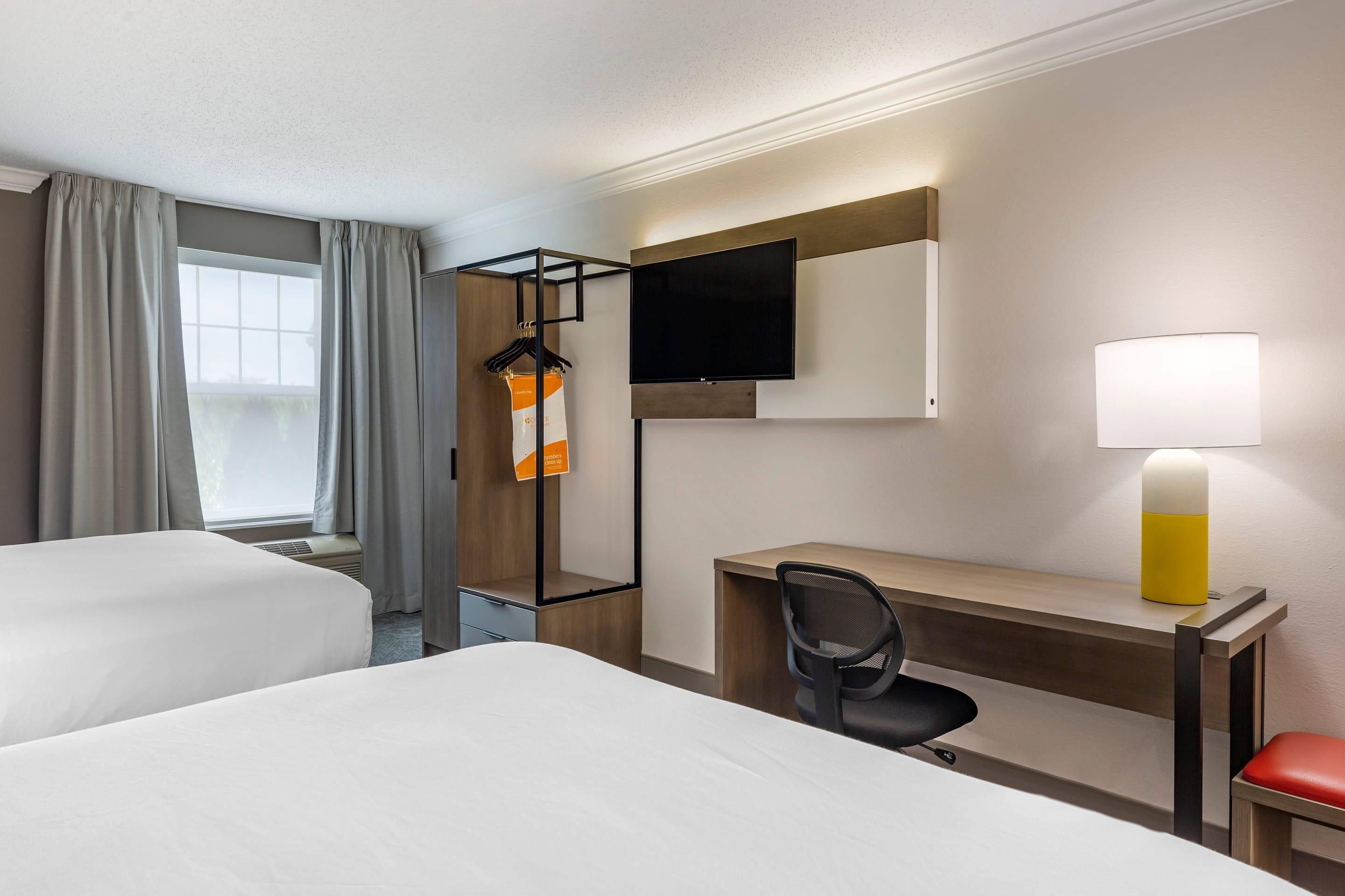A hotel room with two beds, a desk, a chair, a TV, and a lamp.