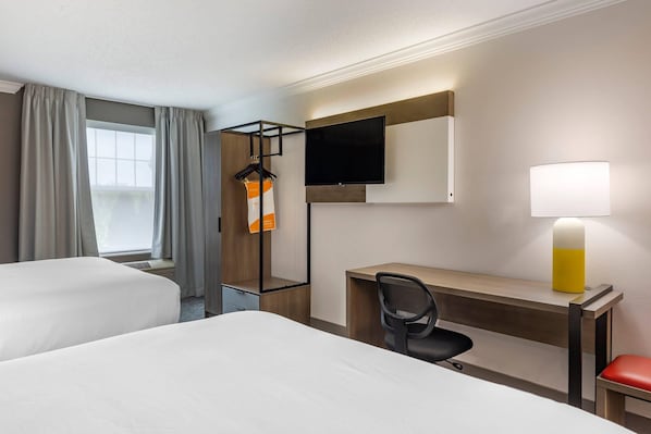 A hotel room with two beds, a desk, a chair, a TV, and a lamp.