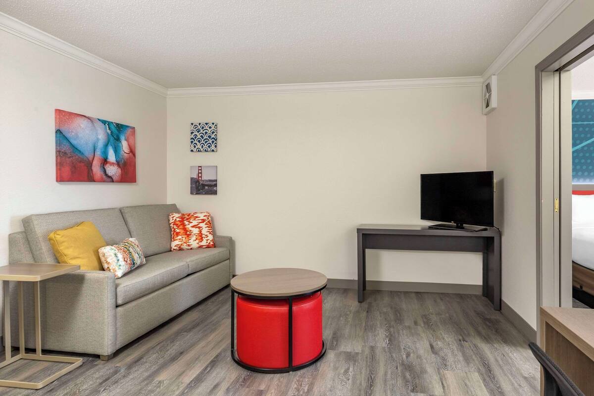 A modern living room with a grey sofa, red accent stool, and a flat-screen TV on a dark table.