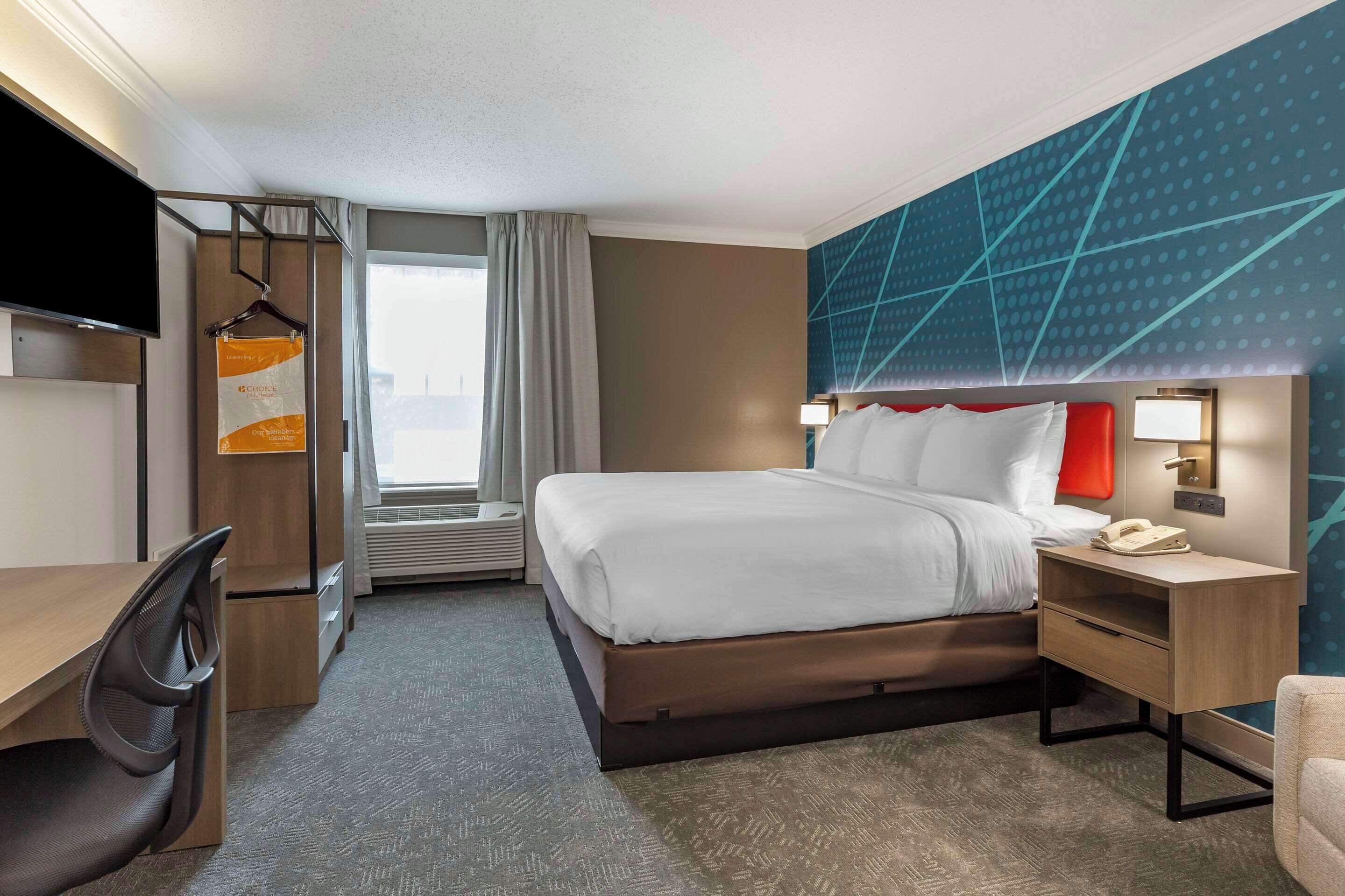 A hotel room with a bed, desk, chair, TV, and a view of the city.