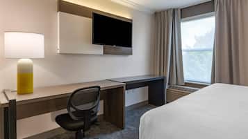 A hotel room with a bed, a desk with a chair, a flat-screen TV, a lamp, and a window with curtains.