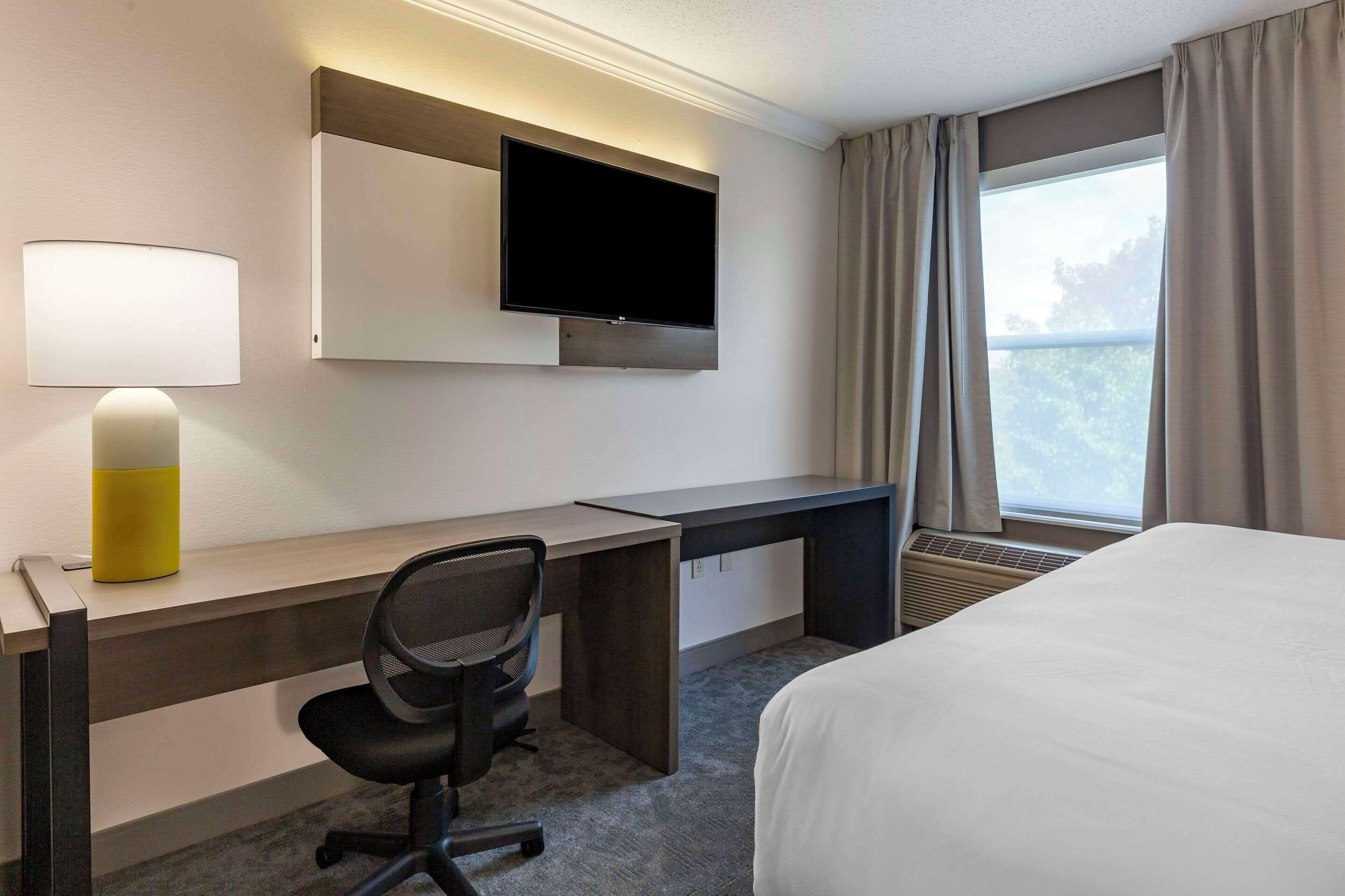 Habitación de hotel con cama, escritorio, silla, televisor, lámpara y ventana con cortinas.