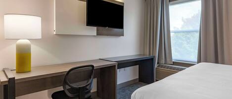 Habitación de hotel con cama, escritorio, silla, televisor, lámpara y ventana con cortinas.