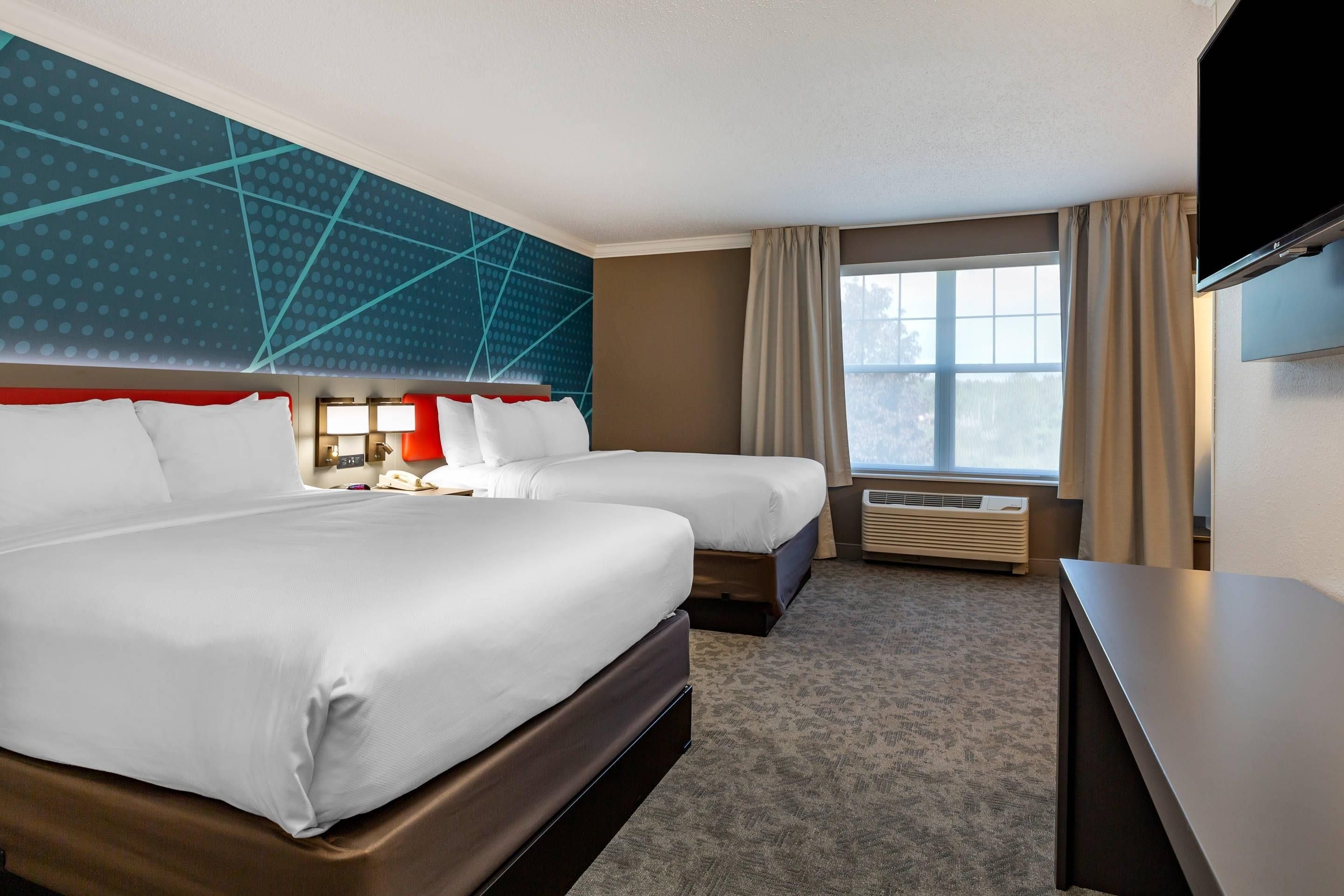 A hotel room with two beds, a TV, a desk, a window with curtains, and a wall with a geometric pattern.