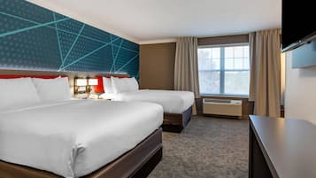 A hotel room with two beds, a TV, a desk, a window with curtains, and a wall with a geometric pattern.