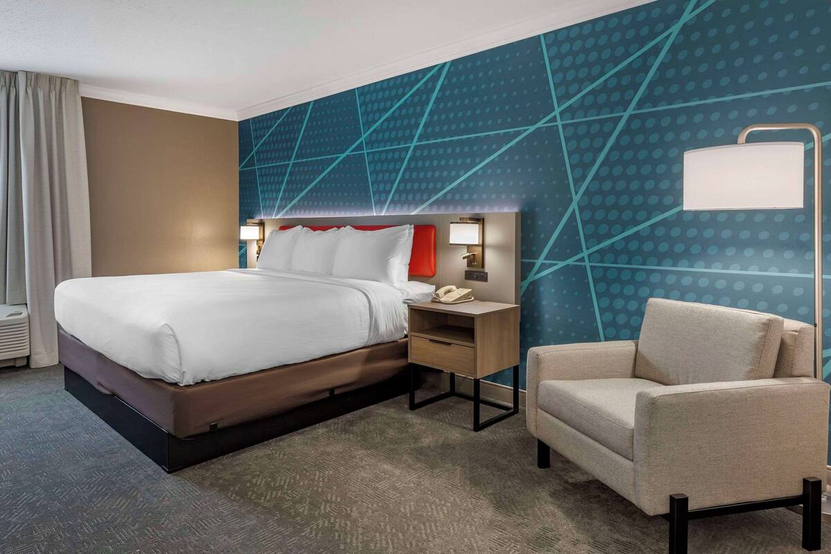 A hotel room with a large bed, a bedside table with a lamp, a chair, and a wall with a geometric pattern.