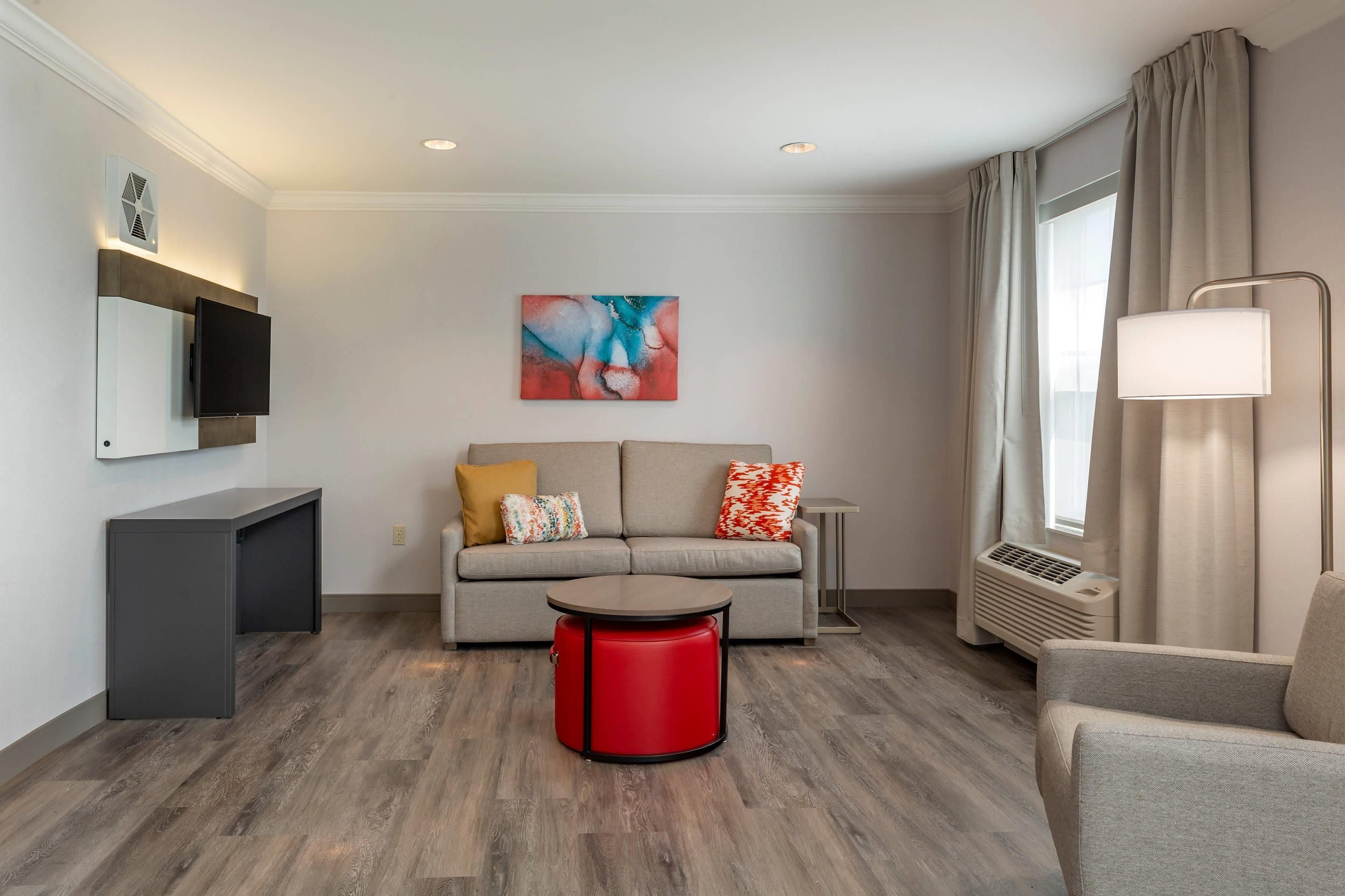 A modern living room with a sofa, a red ottoman, a flat-screen TV, and a painting on the wall.