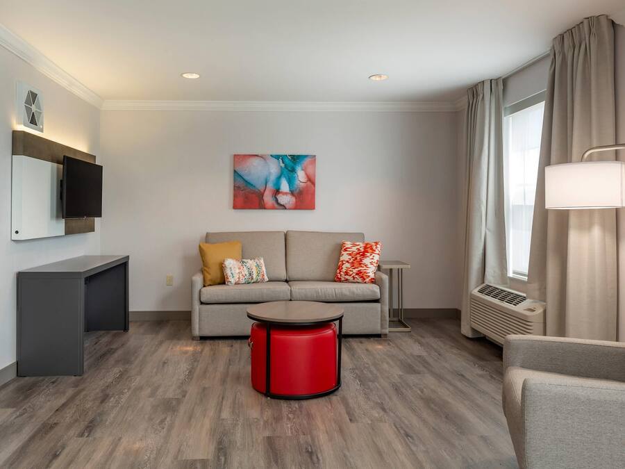 A modern living room with a sofa, a red ottoman, a flat-screen TV, and a painting on the wall.