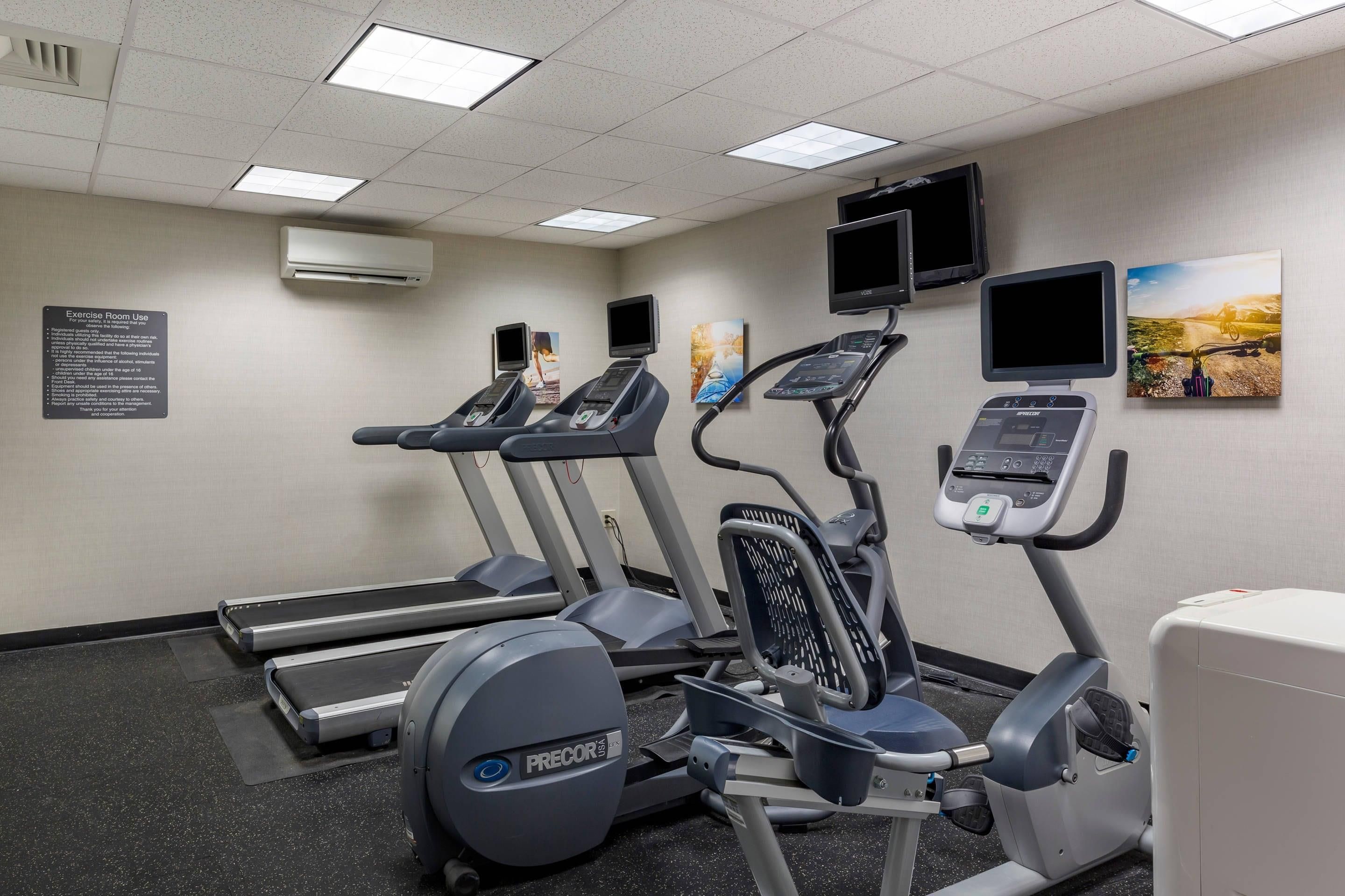 A fitness room with treadmills, an elliptical, and a stationary bike, equipped with digital displays and a wall-mounted TV.