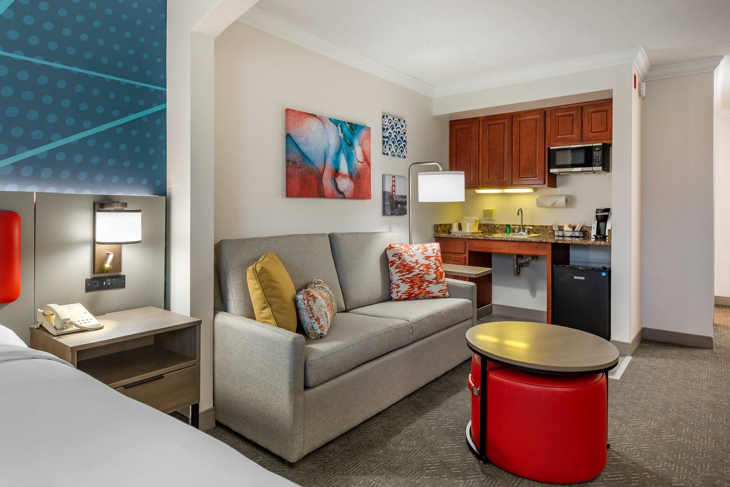 A hotel room with a bed, a desk with a phone, a sofa with cushions, a red ottoman, a kitchenette with a microwave, and a painting on the wall.