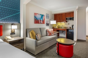 A hotel room with a bed, a desk with a phone, a sofa with cushions, a red ottoman, a kitchenette with a microwave, and a painting on the wall.