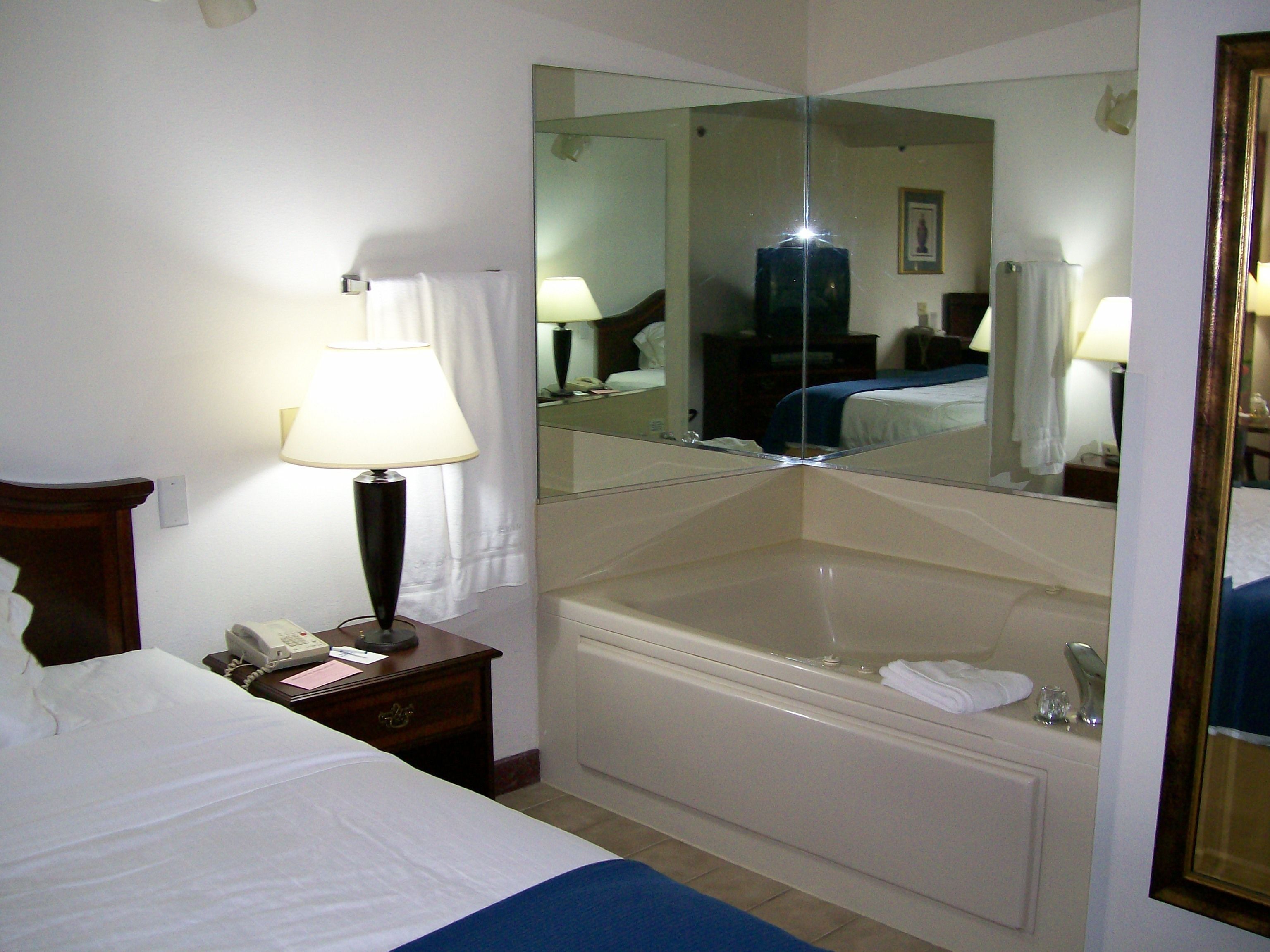 A hotel room with a bed, a bedside table with a lamp, a mirror, and a bathtub.