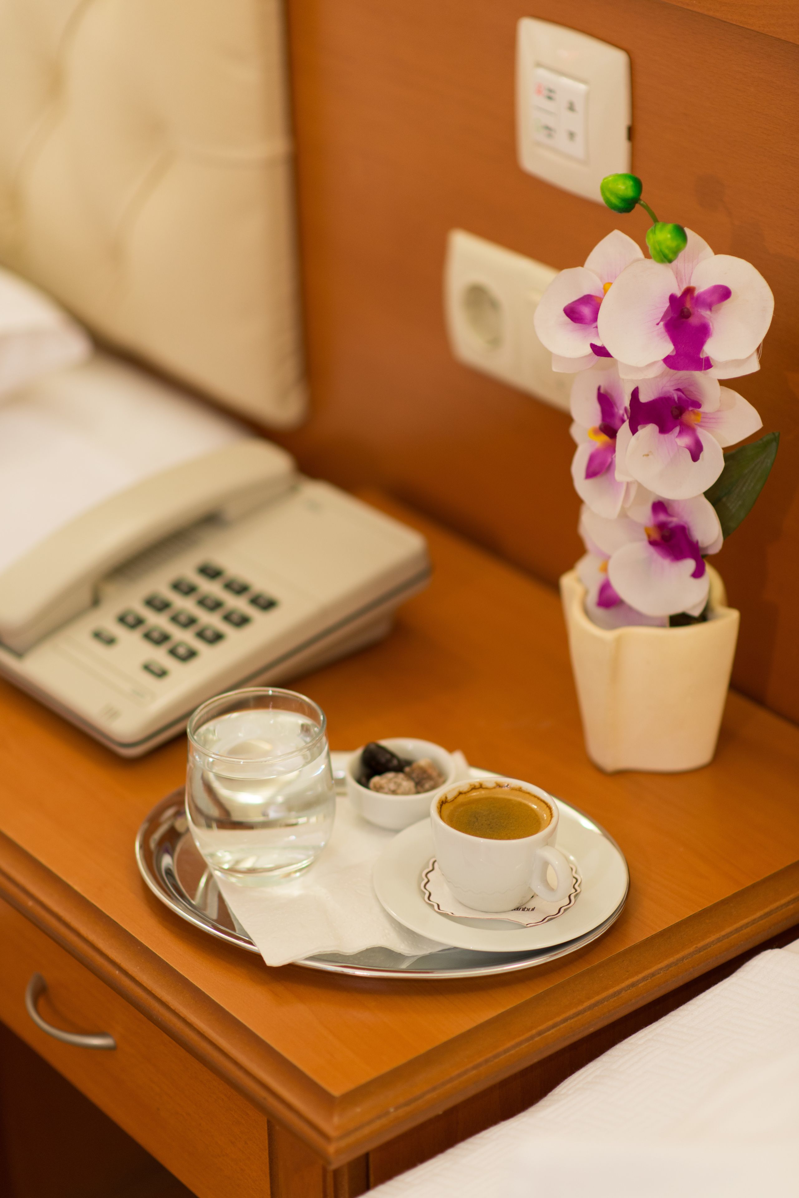 A hotel room with a bedside table featuring a phone, a glass of water, a cup of coffee, and a vase with an orchid.
