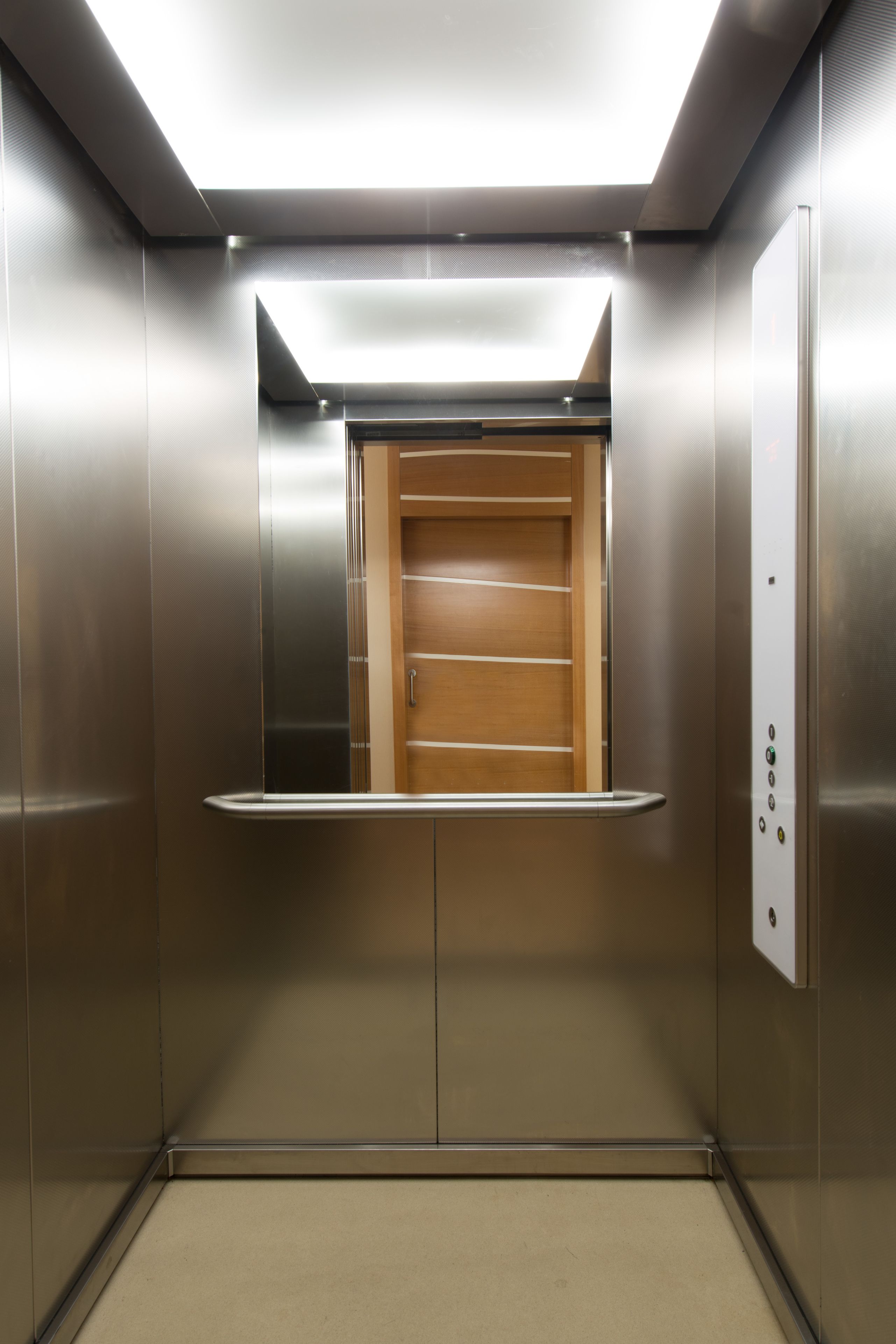 Interior de un ascensor con paredes metálicas, una puerta de madera y un panel de control a la derecha.
