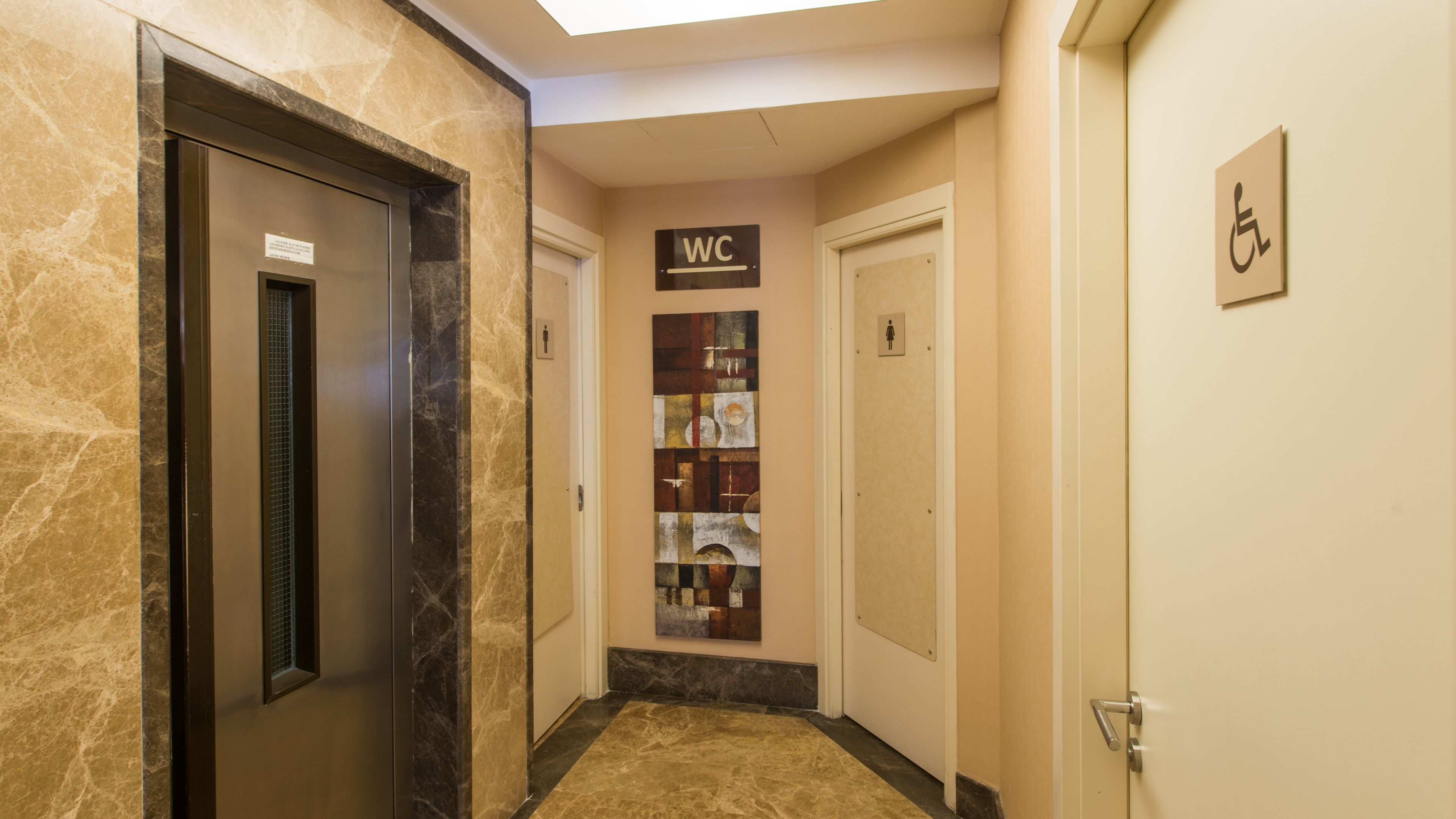 Un couloir avec un sol en marbre, un ascenseur, un panneau indiquant les toilettes et un panneau indiquant l’accès pour les personnes handicapées.