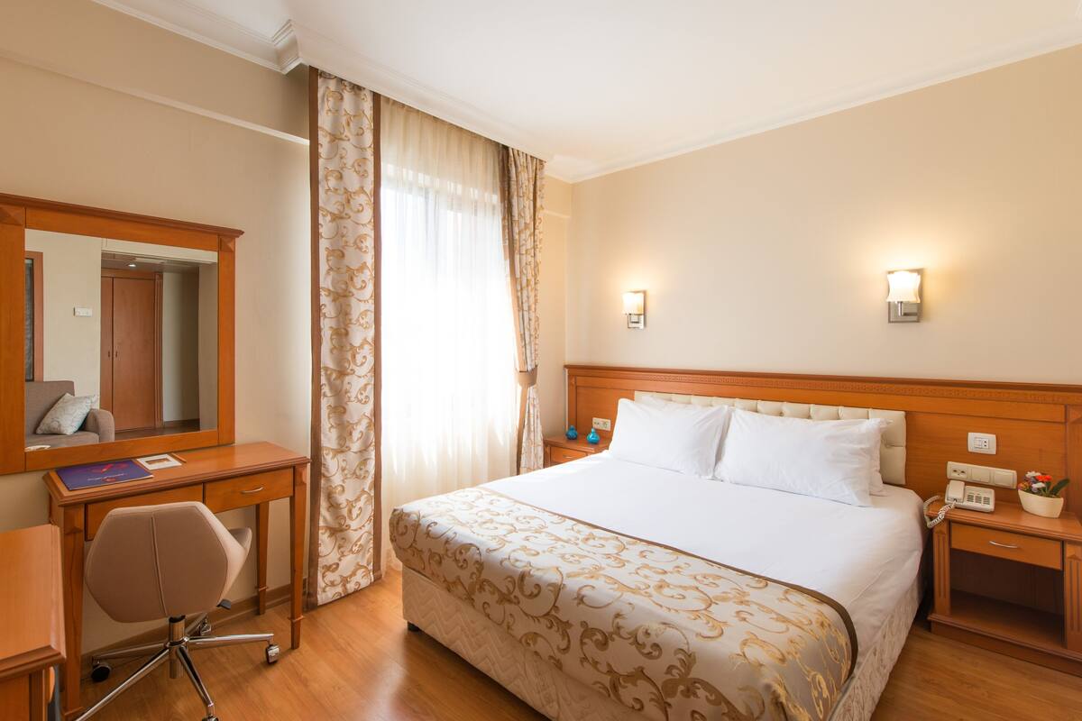 A hotel room with a bed, a desk, a chair, a mirror, a nightstand, and a lamp.