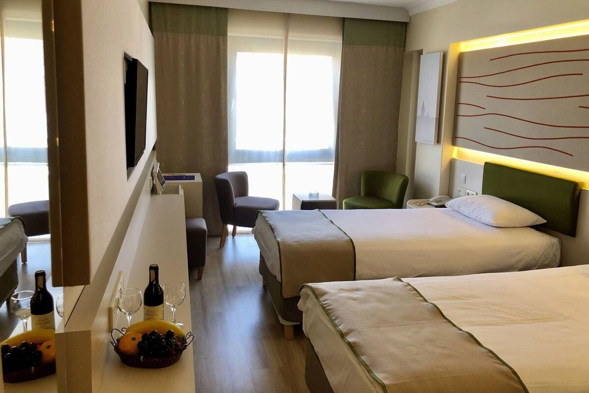 A hotel room with two beds, a desk with a bottle of wine, a chair, a television, and a large window.