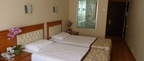 A hotel room with three single beds, a bedside table with a phone, a small table with a vase of flowers, and a mirror on the wall.