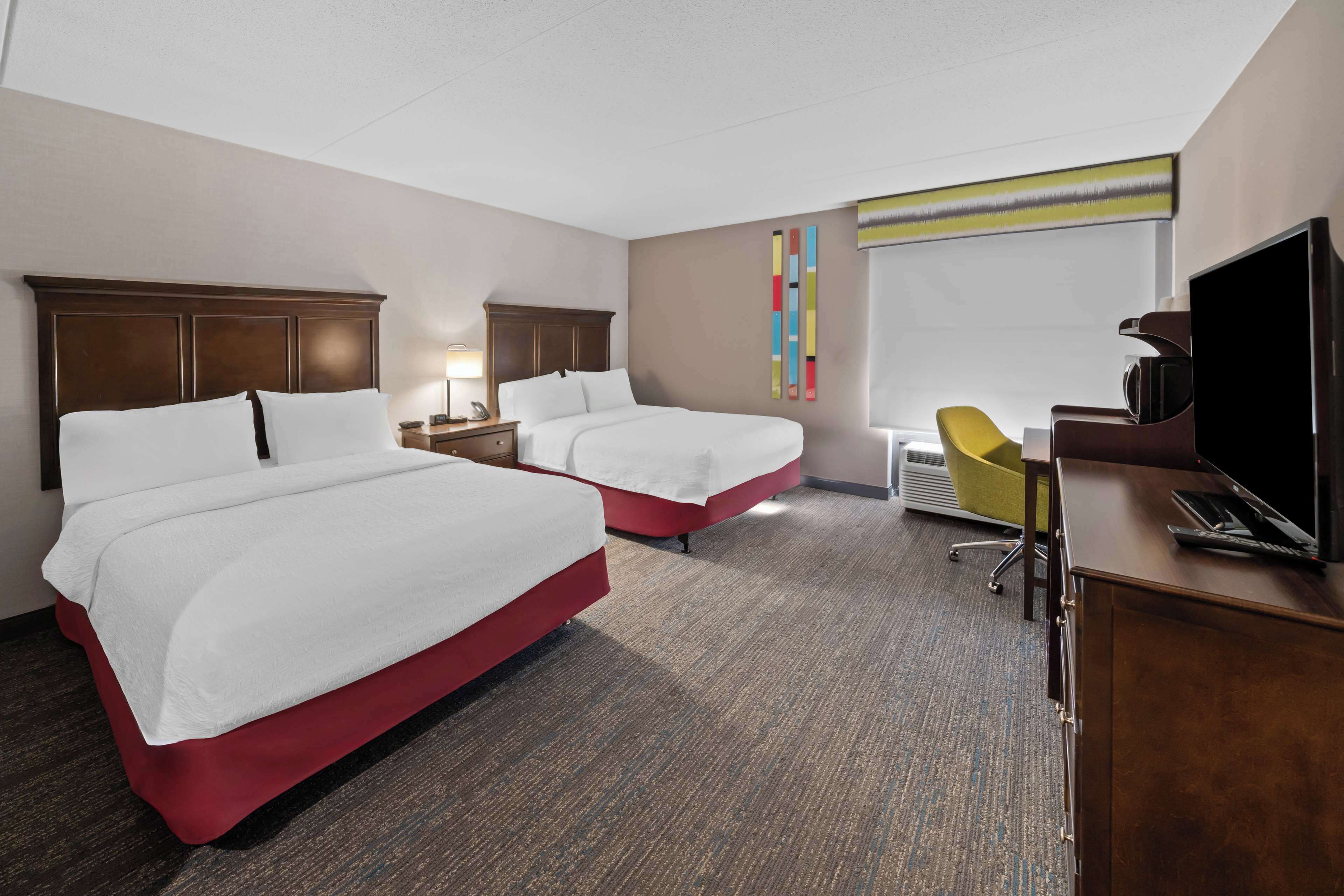 Une chambre d’hôtel avec deux lits, un bureau avec une télévision, une chaise et une œuvre d’art colorée au mur.