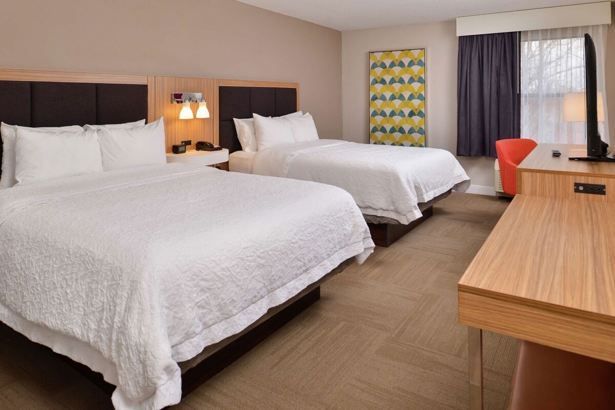 Une chambre d’hôtel avec deux lits, un bureau, une chaise, une table de chevet, une lampe et une œuvre d’art murale.
